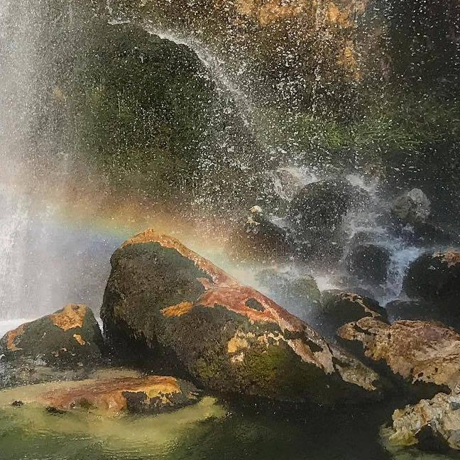 a rainbow in the white spray of a waterfall, lit by sunlight, over brown rocks. in the bottom left corner, there is sandy yellow-green water. the brown rocks in the bottom right corner are in shadow.